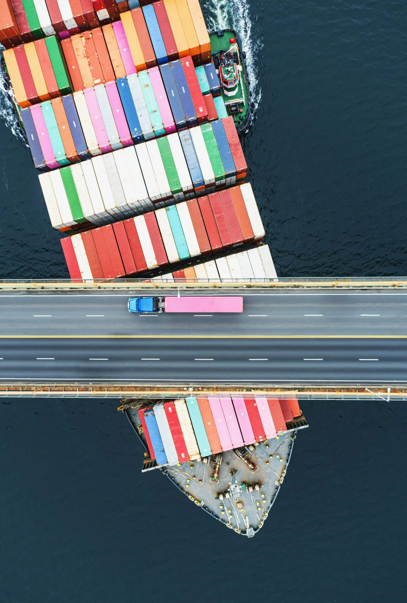 An aerial view of a container ship passing under a bridge, a truck crossing above and a second ship below