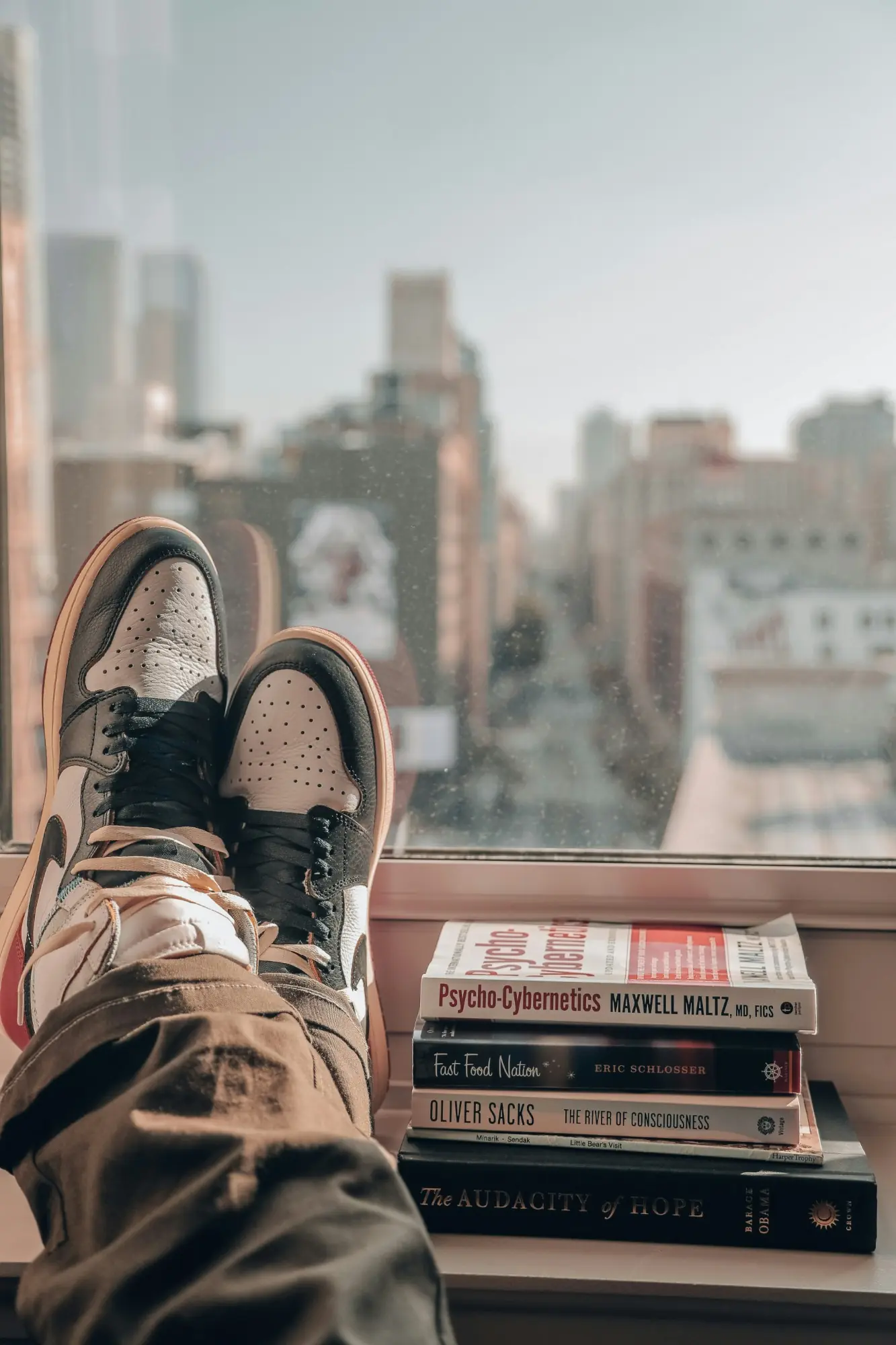 Sneakered feet resting on a window ledge beside a stack of books, overlooking a city street