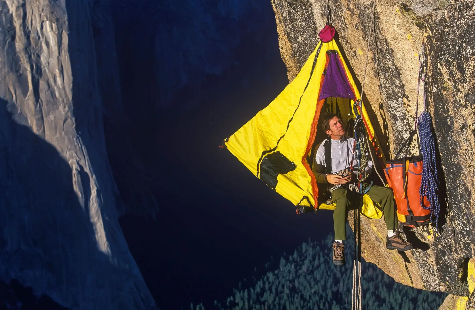 A climber resting in a yellow portaledge tent on a granite cliff