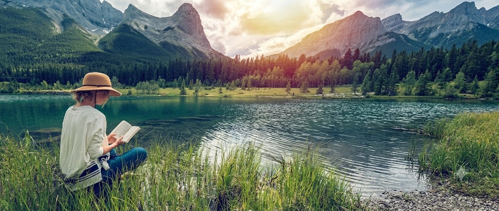 A woman sitting by a mountain lake, reading an open book outdoors.